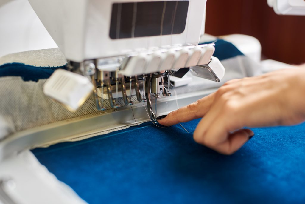 Close-up of woman hand at computerized embroidery machine on background of bright blue textile detail. Specially engineered machine with multi-needle fixed embroidery head.